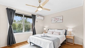 A bedroom with a bed, bedside tables, a ceiling fan, a window with curtains, and a wall-mounted air conditioner. - Retro Retreat - Mornington (Mornington)