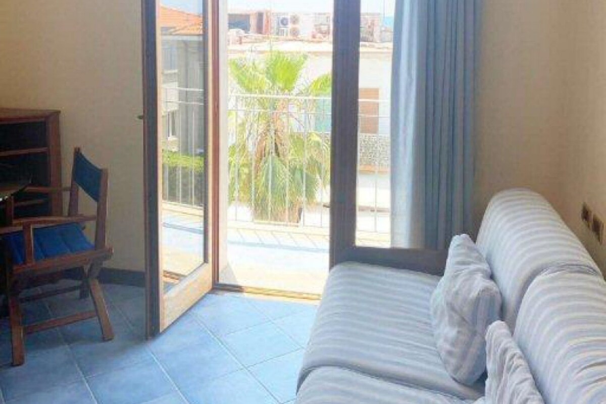 A room with a bed, a desk, and a chair. There is a sliding glass door leading to an outdoor area with a view of buildings and greenery.