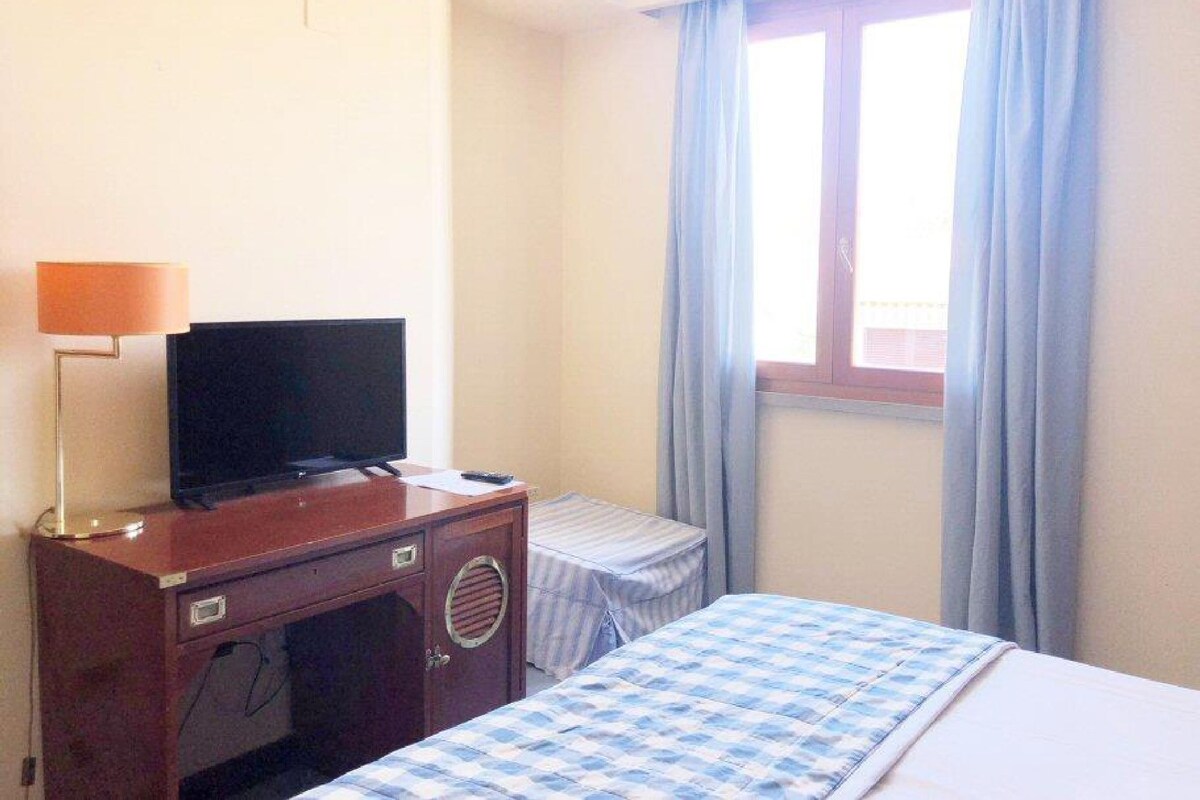 A hotel room with a bed, a desk with a television, a window with curtains, and a small table with a chair.
