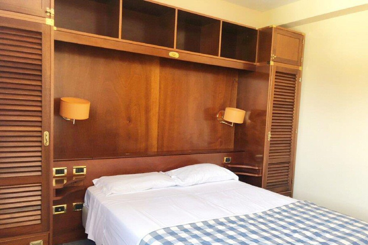 A hotel room with a wooden headboard, two bedside lamps, a bed with white linens, and a checkered throw blanket.