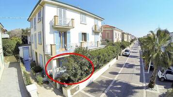 Un edificio di due piani con un balcone, un piccolo giardino e vista sulla strada con auto parcheggiate e palme.