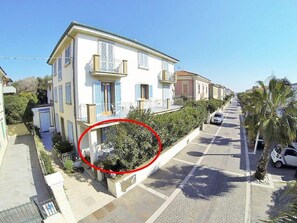 A two-story building with a balcony, a small garden, and a street view with parked cars and palm trees. - Zì Maria (Cecina)