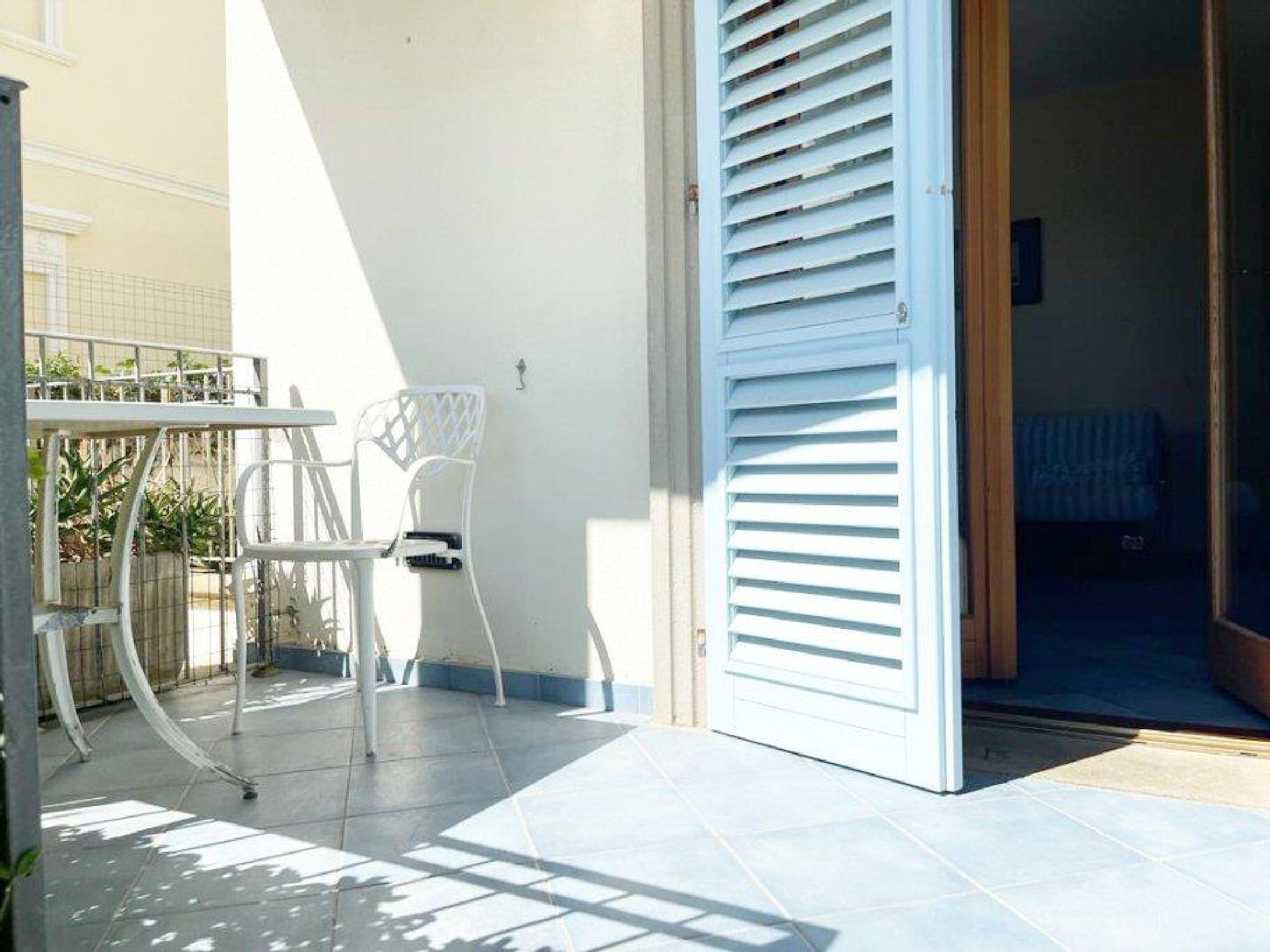 Eine Terrasse mit einem weißen Metalltisch und -stühlen, einer weißen Tür mit horizontalen Rollläden und einem gefliesten Boden.
