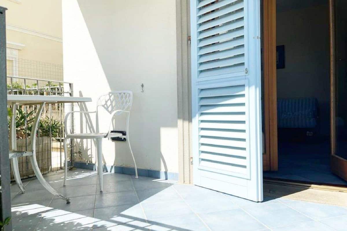Un espace patio avec une table et des chaises en métal blanc, une porte blanche dotée de volets à lames horizontales et un sol carrelé.