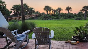 A patio with two Adirondack chairs, a wooden deck, and a view of a grassy area with palm trees and a wooden bridge. - Lovely and affordable oceanfront retreat, just steps from St. Augustine Beach (St. Augustine)