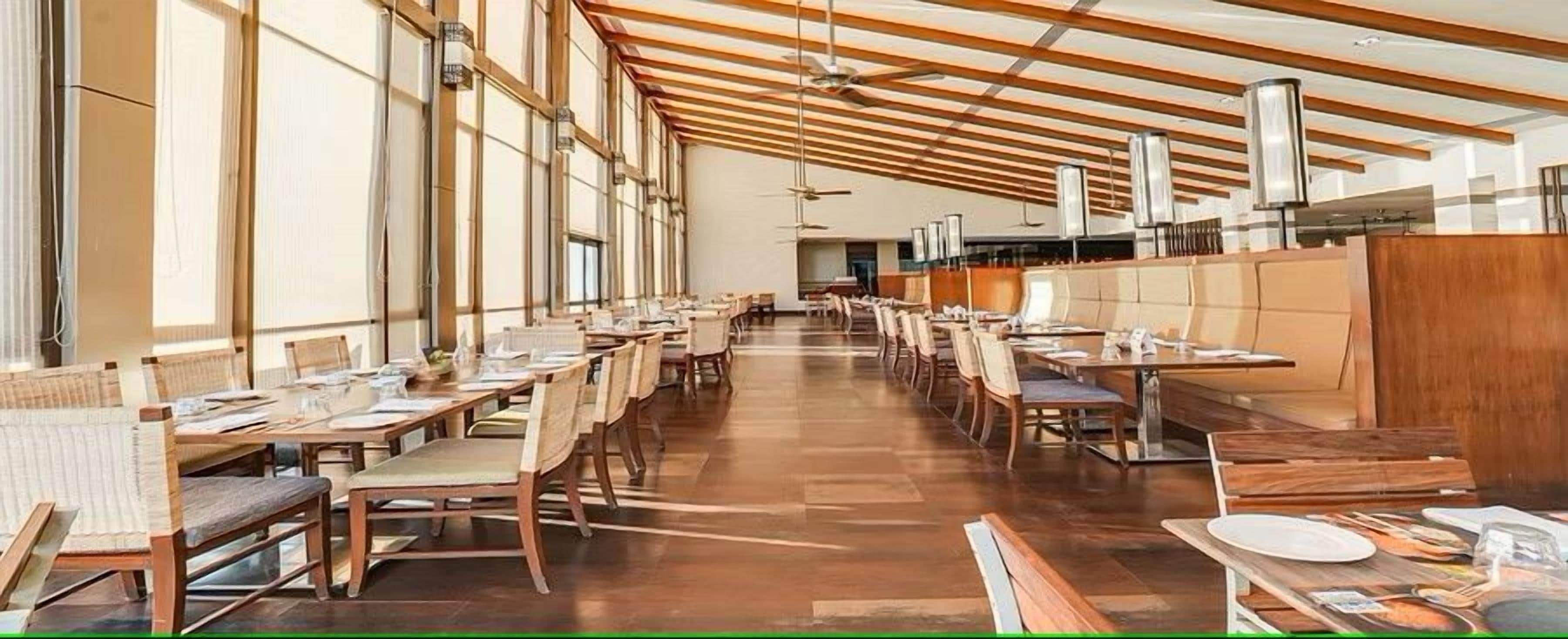 A spacious dining area with long tables and chairs, large windows, and a high ceiling with exposed beams.