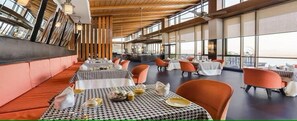 En restaurang med högt i tak, stora fönster och orange stolar. Borden är dukade med vita dukar och svarta och vita rutiga bordstabletter.