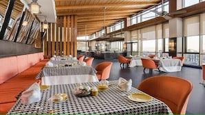 A restaurant with high ceilings, large windows, and orange chairs. Tables are set with white tablecloths and black and white checkered table runners. - Club Mahindra Resort - Tungi (Ootacamund)