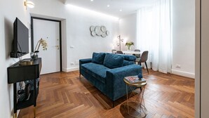 Ein modernes Wohnzimmer mit einem blauen Sofa, einem kleinen runden Tisch, einem Fernseher auf einem Schrank und einem Essbereich im Hintergrund.
