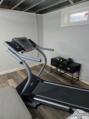 Een loopband in een kamer met een houten vloer, een klein zwart tafeltje met trainingsballen, en een raam met een bord.