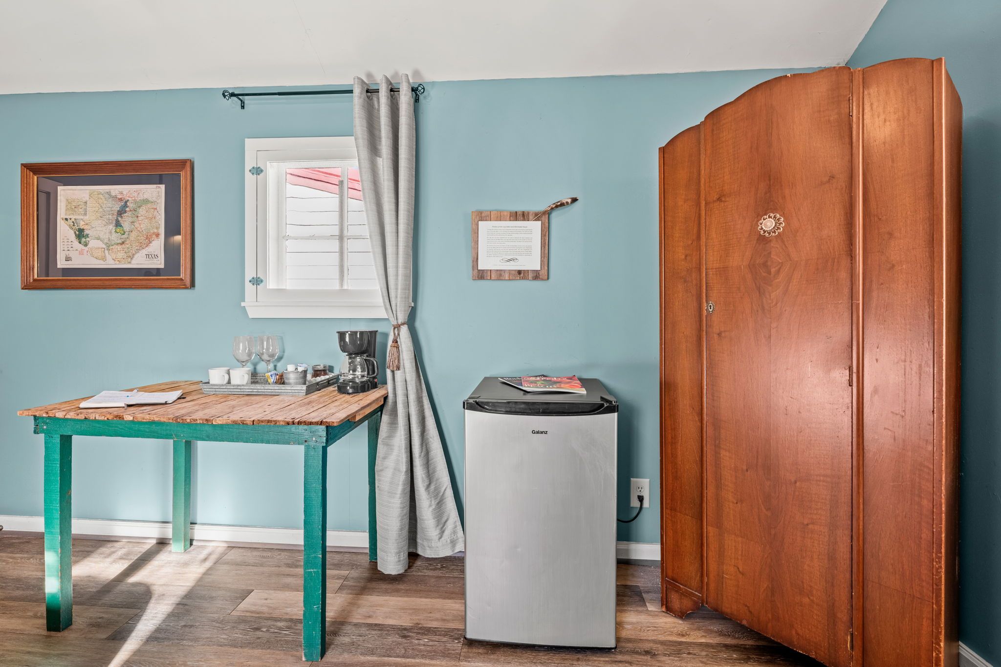 A room with a wooden cabinet, a small table with a coffee maker, a framed map, and a window with curtains.