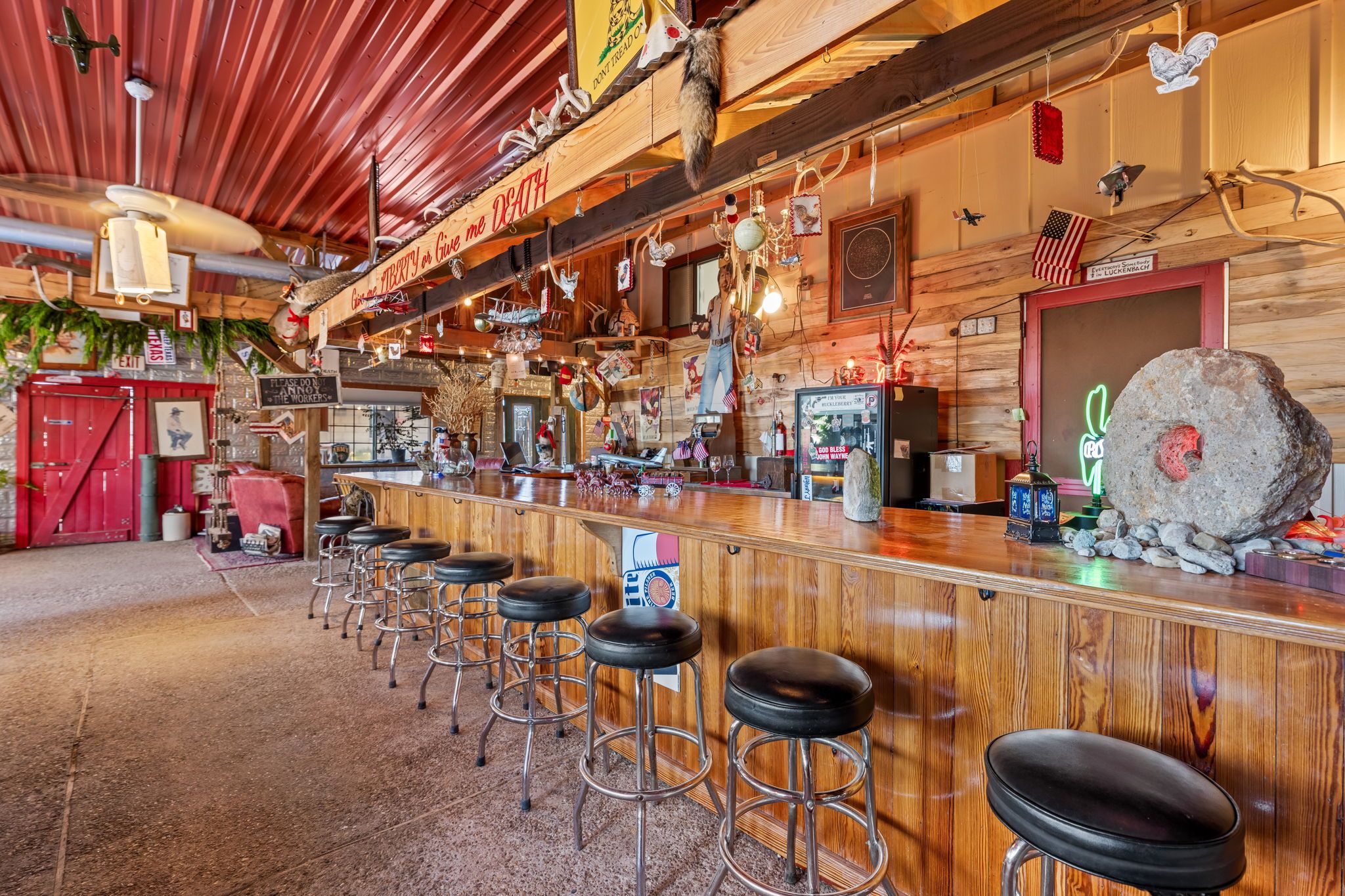 A rustic bar area with wooden bar stools, a neon sign, and various decorations including a large rock and American flags.