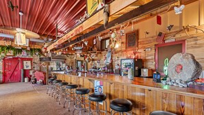 A rustic bar area with wooden bar stools, a neon sign, and various decorations including a large rock and American flags.