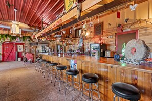 Un coin bar au style rustique, avec des tabourets de bar en bois, un panneau néon et diverses décorations, notamment un gros rocher et des drapeaux américains.