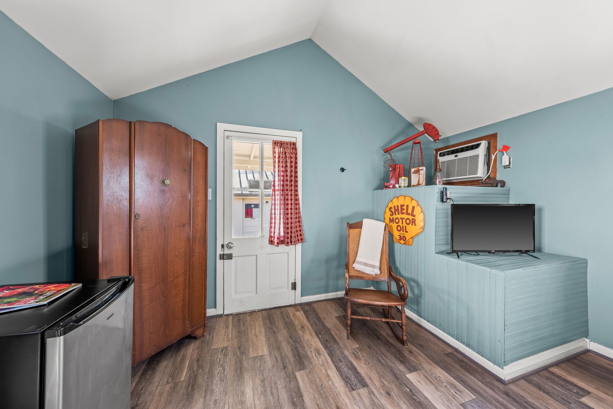 A room with a wooden wardrobe, a white door with a red checkered curtain, a vintage Shell sign, a wicker chair, and a television on a blue cabinet.