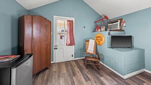A room with a wooden wardrobe, a white door with a red checkered curtain, a vintage Shell sign, a wicker chair, and a television on a blue cabinet.