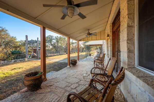 A covered outdoor area with stone flooring, wooden chairs, a ceiling fan, and a rustic stone wall.