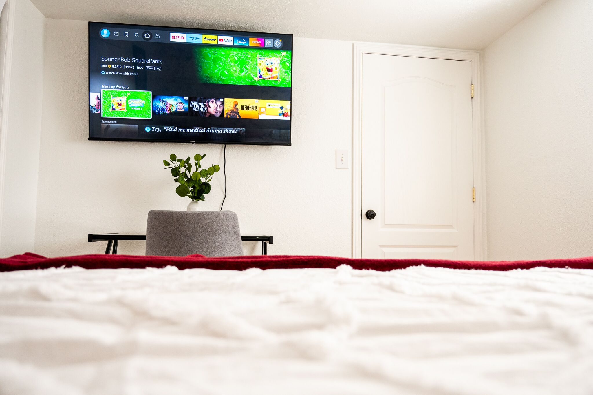 Un dormitorio con una cama, un televisor montado en la pared, una planta pequeña sobre una mesa y una puerta blanca.