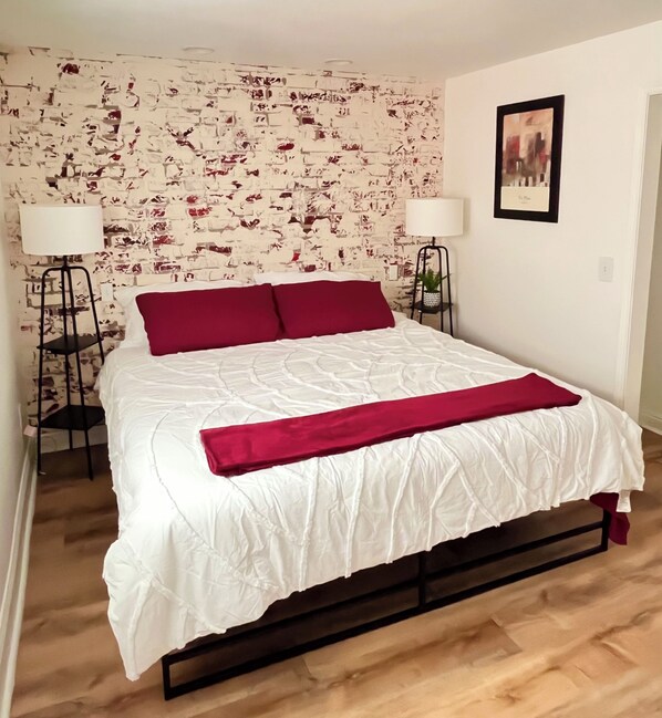 A neatly made bed with a white comforter and a red throw blanket, positioned in a room with a brick-patterned wall and a framed picture on the wall. - Private Entrance\Modern&Cozy Central Bliss\King Villa\55" 4k Roku\Full Kitchen (Colorado Springs)
