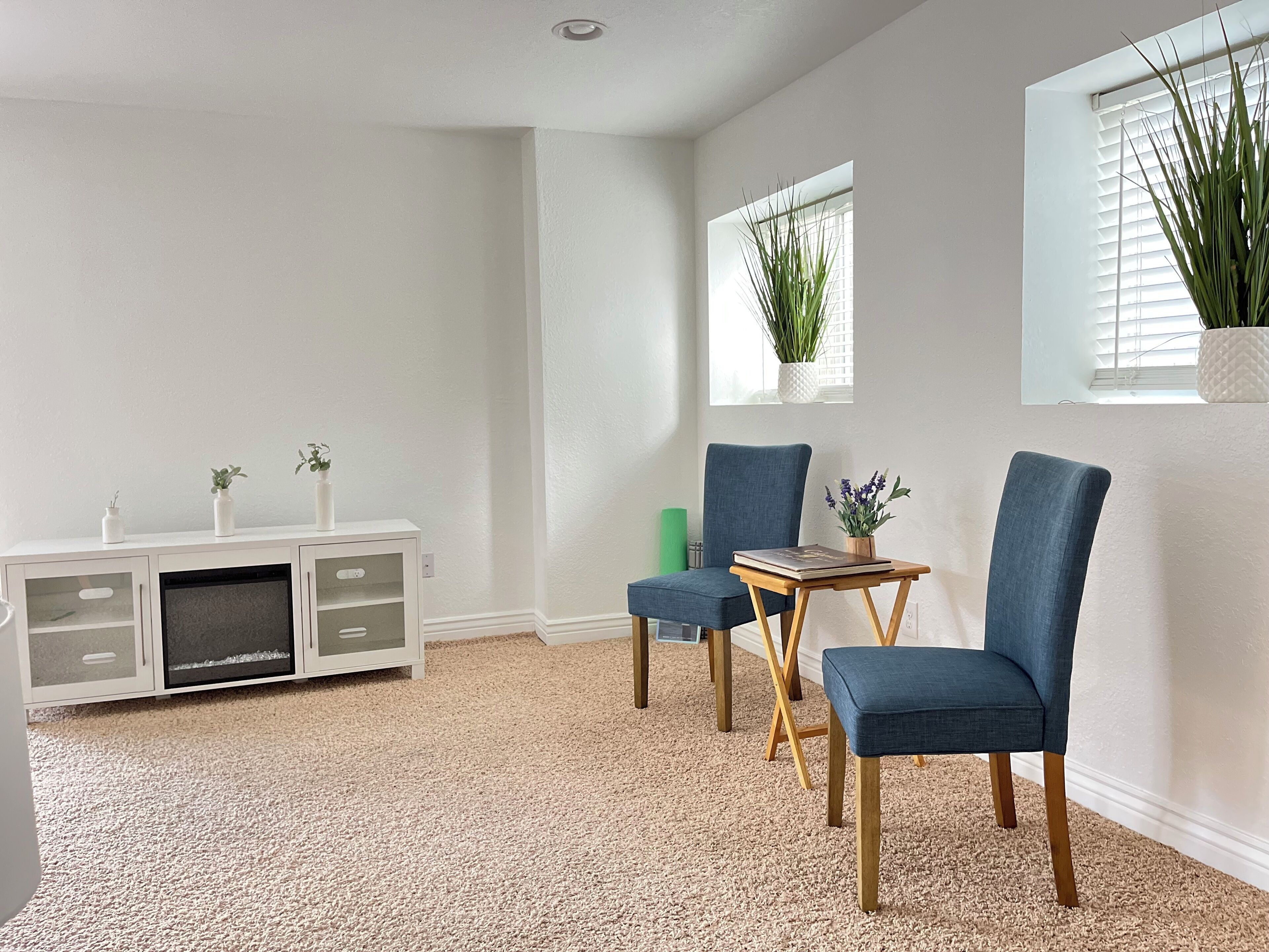 Una habitación con alfombra blanca, un armario blanco con puertas de cristal, dos sillas azules, una pequeña mesa de madera y una ventana con una planta.