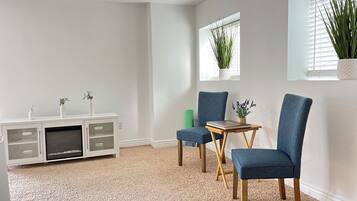 Una habitación con alfombra blanca, un armario blanco con puertas de cristal, dos sillas azules, una pequeña mesa de madera y una ventana con una planta.