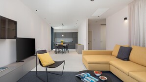 Ein modernes Wohnzimmer mit einer Couch, einem Couchtisch und einem Fernseher. Im Hintergrund befindet sich ein Essbereich mit Tisch und Stühlen.