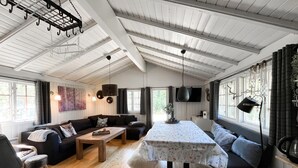 Un salon chaleureux avec un canapé, une table basse et une table à manger recouverte d’une nappe à motifs floraux. On y trouve des fenêtres avec des rideaux, un luminaire suspendu et un téléviseur fixé au mur.