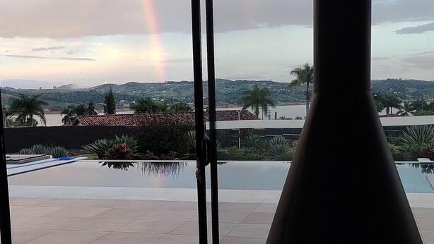 Un interior moderno con un amplio ventanal que da a una piscina y un paisaje pintoresco, incluyendo un arcoíris.