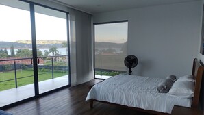 Un dormitorio con un amplio ventanal corredero que da a un balcón, una cama con ropa de cama blanca, un ventilador de pie y un cabecero de madera.