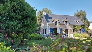 Ein Steinhaus mit geneigtem Dach, ein Garten mit verschiedenen Pflanzen und Blumen sowie Sitzgelegenheiten im Freien.