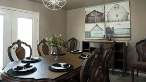 Une salle à manger avec une table en bois dressée pour quatre personnes, entourée de chaises en bois. Un lustre est suspendu au-dessus de la table, et un vase rempli de fleurs y est posé. Les murs sont décorés de tableaux encadrés représentant des granges.