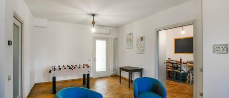 A spacious living area with a foosball table, two blue armchairs, a dining table with chairs, and a kitchen visible through an open door.