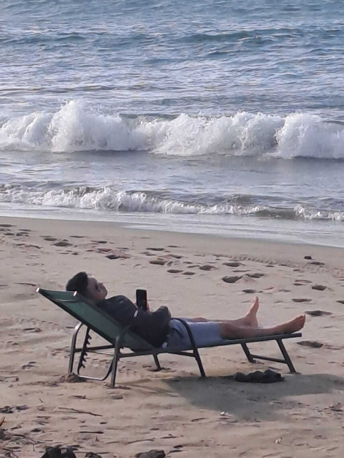 A person lying on a beach chair facing the ocean, with waves crashing onto the shore.