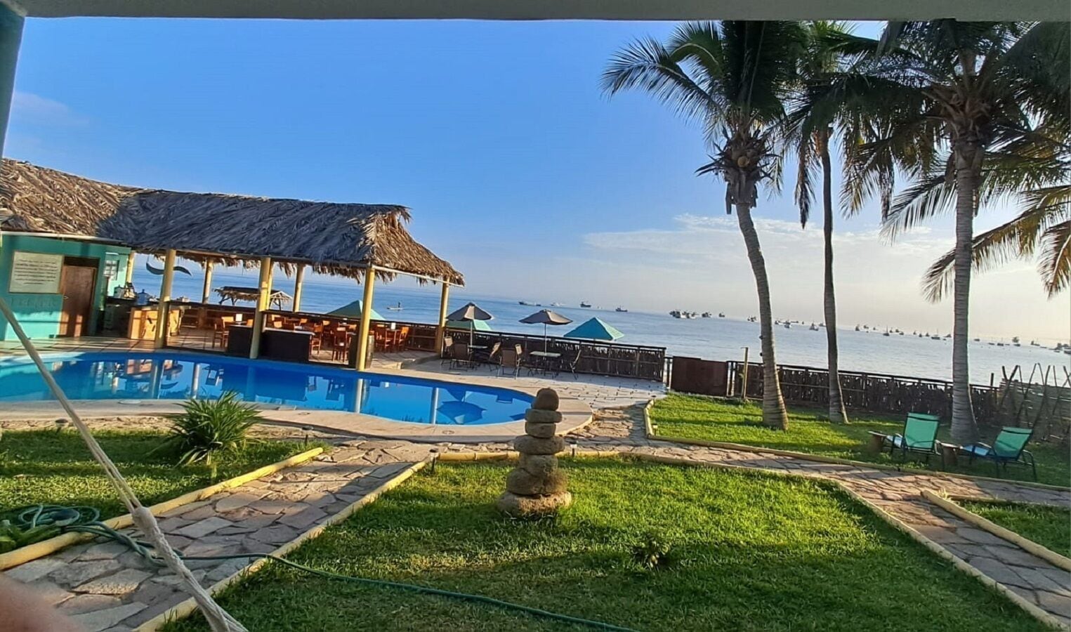 A pool area with a thatched-roof structure, a view of the sea, and palm trees.