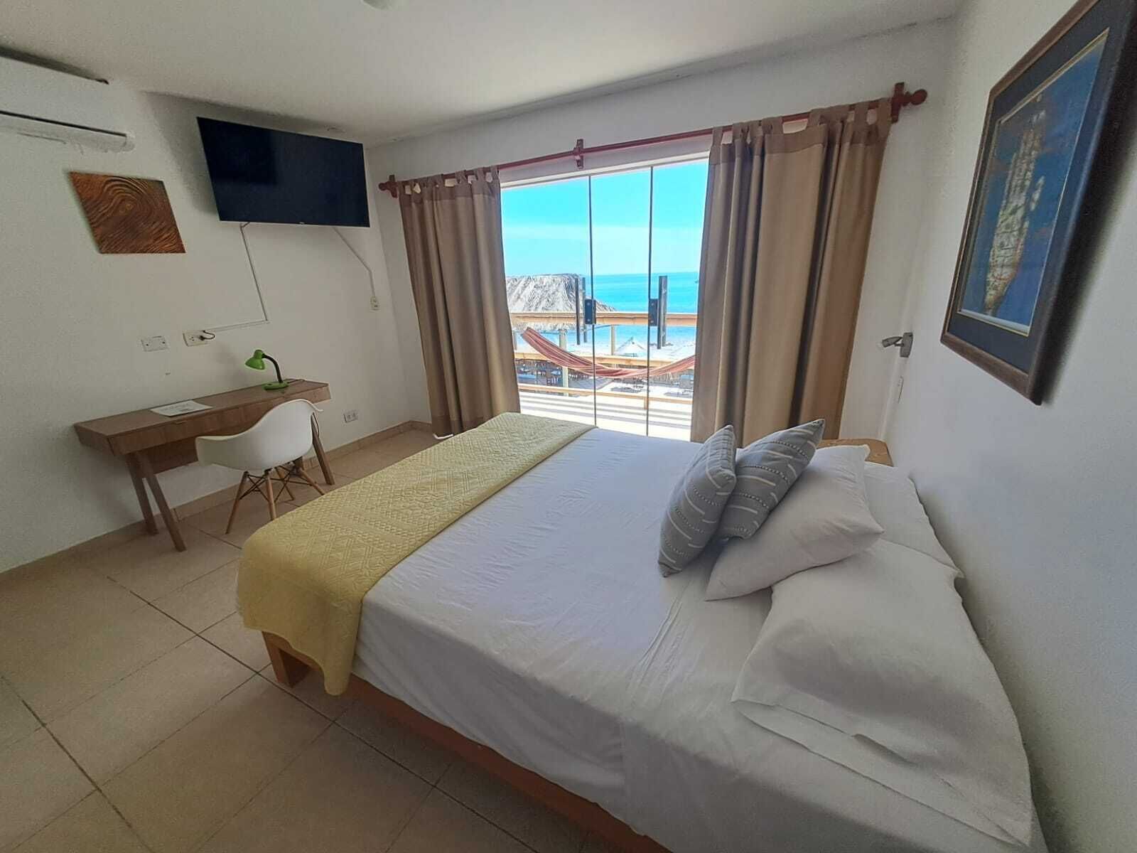 A hotel room with a bed, a desk, a chair, a TV mounted on the wall, and a view of the beach through the window.