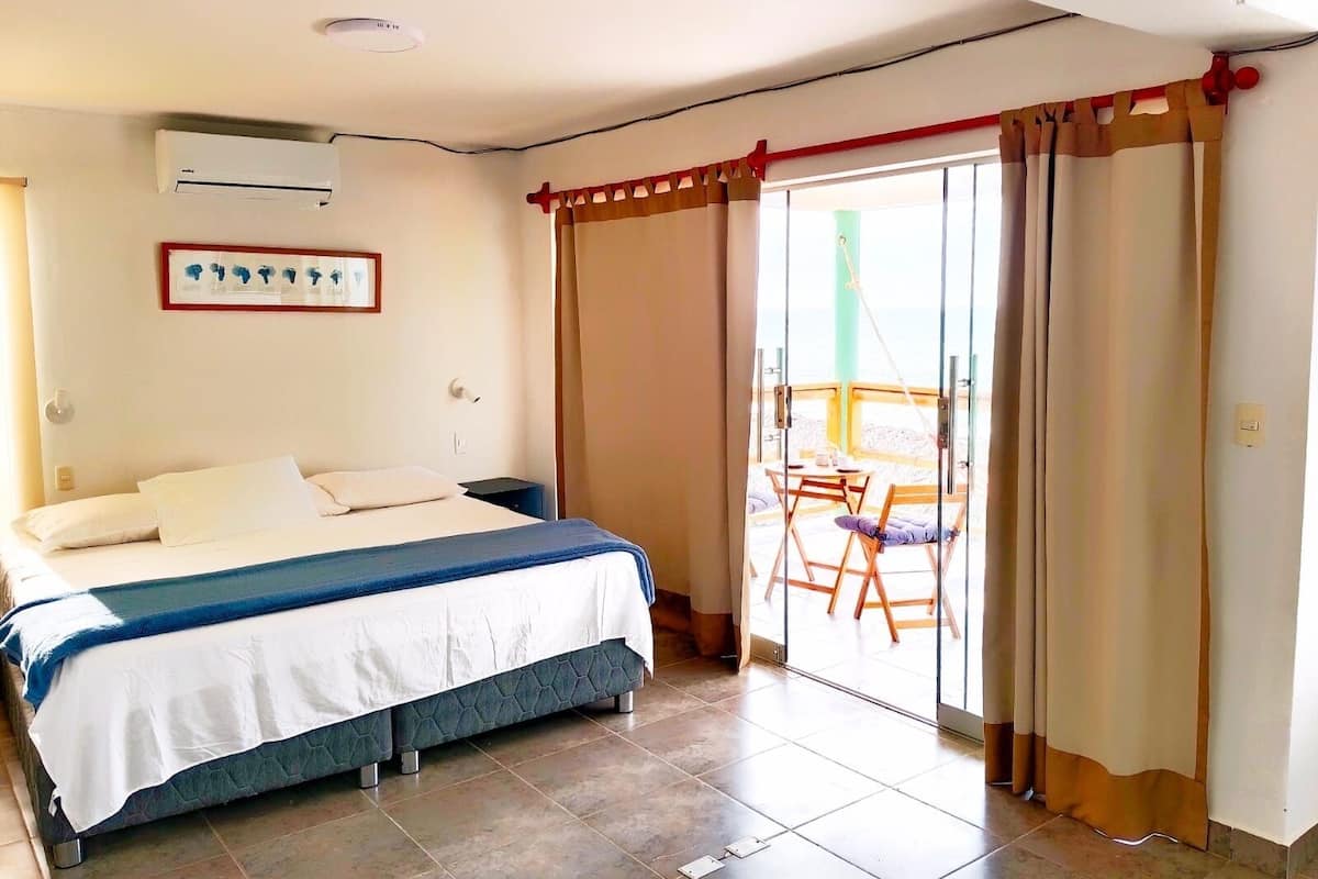 A bedroom with a bed, a small table, and a chair, a sliding glass door to a balcony, and an air conditioning unit on the wall.