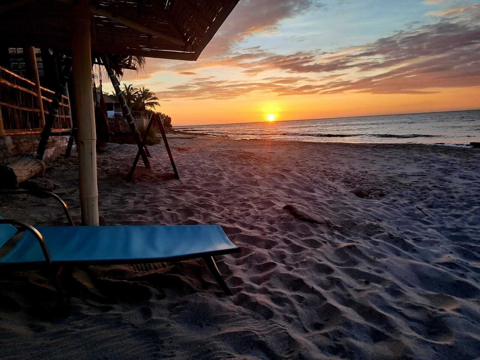 Escena de playa con un atardecer, una estructura de paja y una silla de descanso azul.