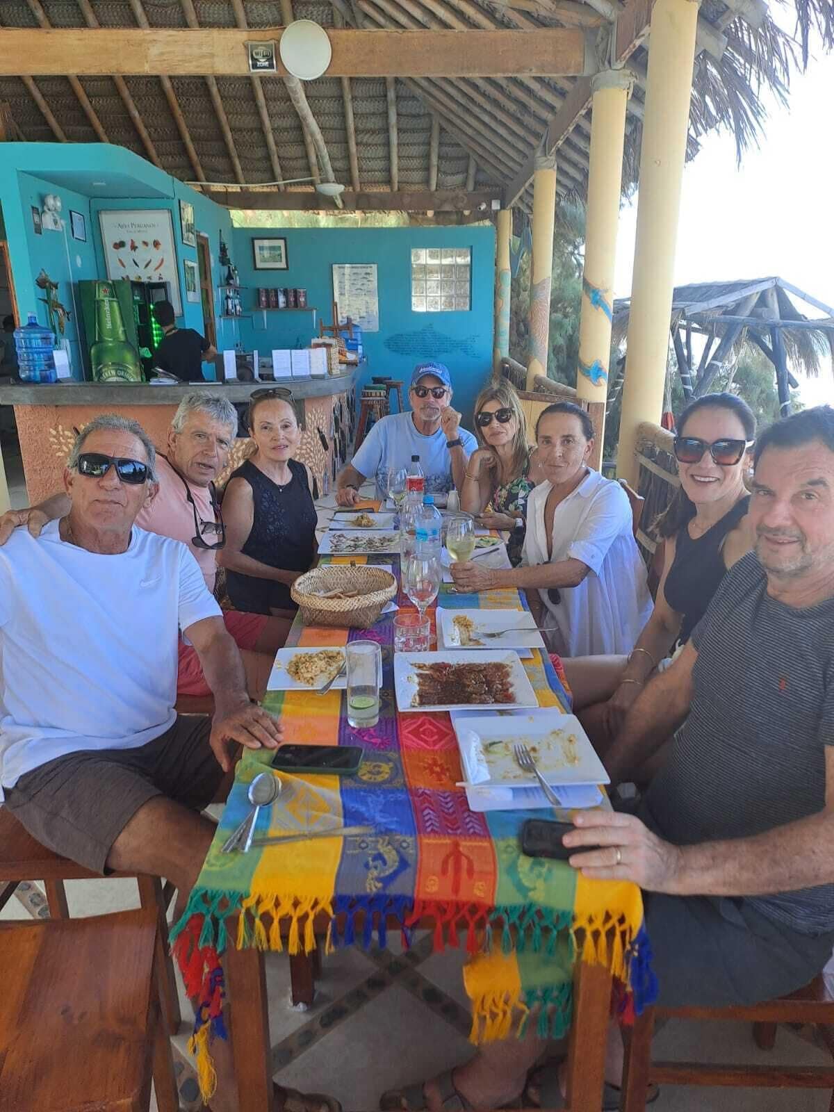 Un groupe de personnes est assis autour d’une table recouverte d’une nappe colorée, sous une structure dont le toit est fait de chaume.