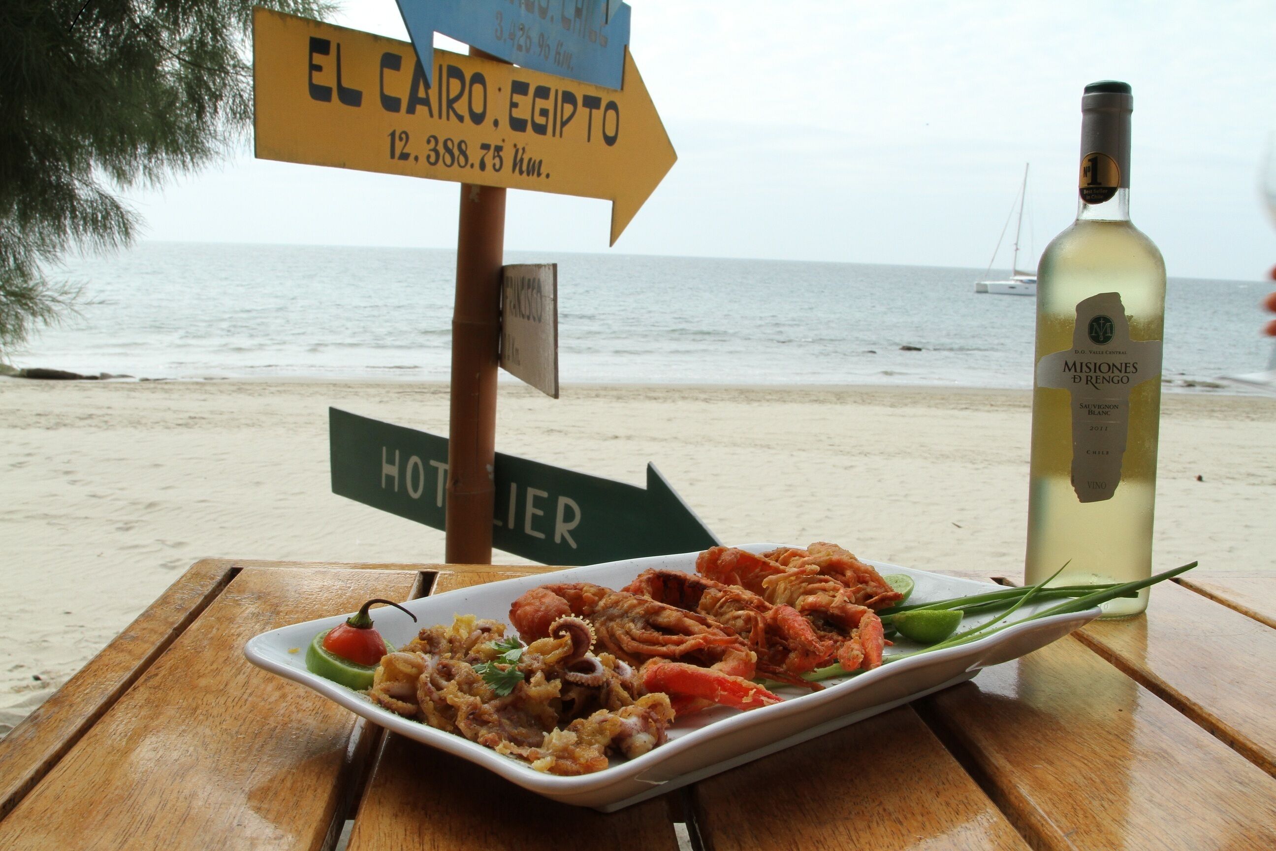 Una mesa con un plato de mariscos y una botella de vino, un cartel con indicaciones y vista a la playa y al mar.