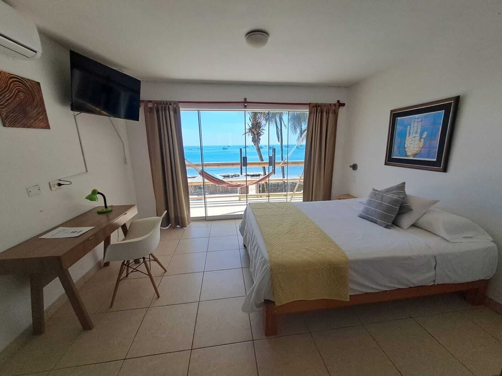 A hotel room with a bed, a desk, a chair, a TV, and a view of the beach and ocean.