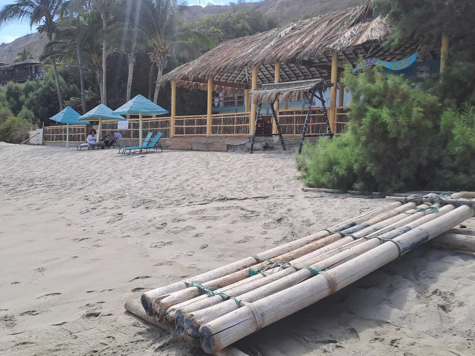 Una cabaña frente al mar con techo de paja, una zona de estar con sillas y una balsa de bambú sobre la arena.