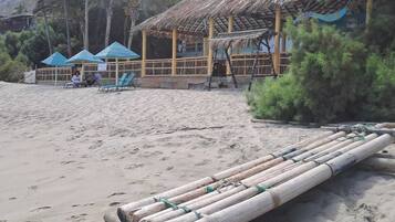 Una cabaña frente al mar con techo de paja, una zona de estar con sillas y una balsa de bambú sobre la arena.