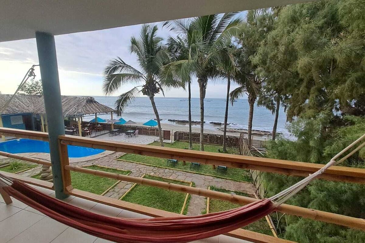 A hammock on a balcony overlooking a pool and beach area with palm trees and a view of the ocean.