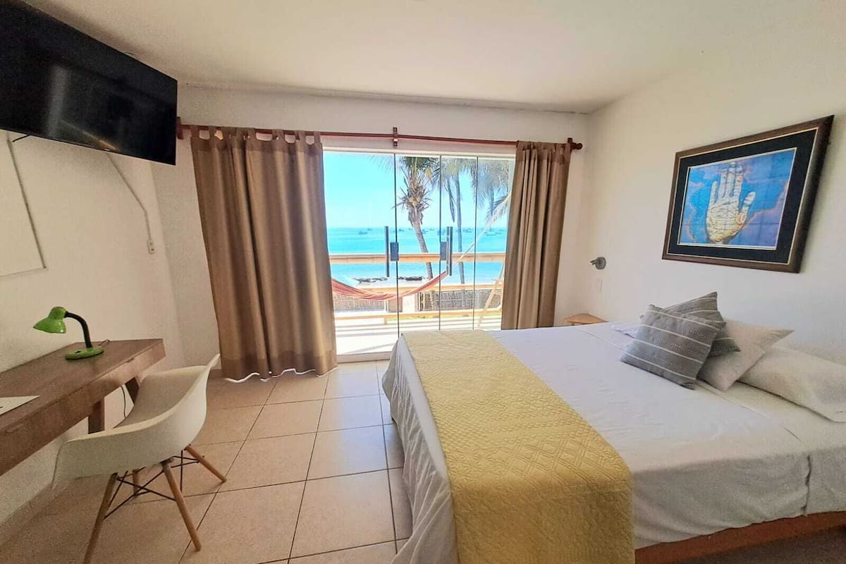 A hotel room with a bed, a desk, a chair, a TV, and a view of the beach.