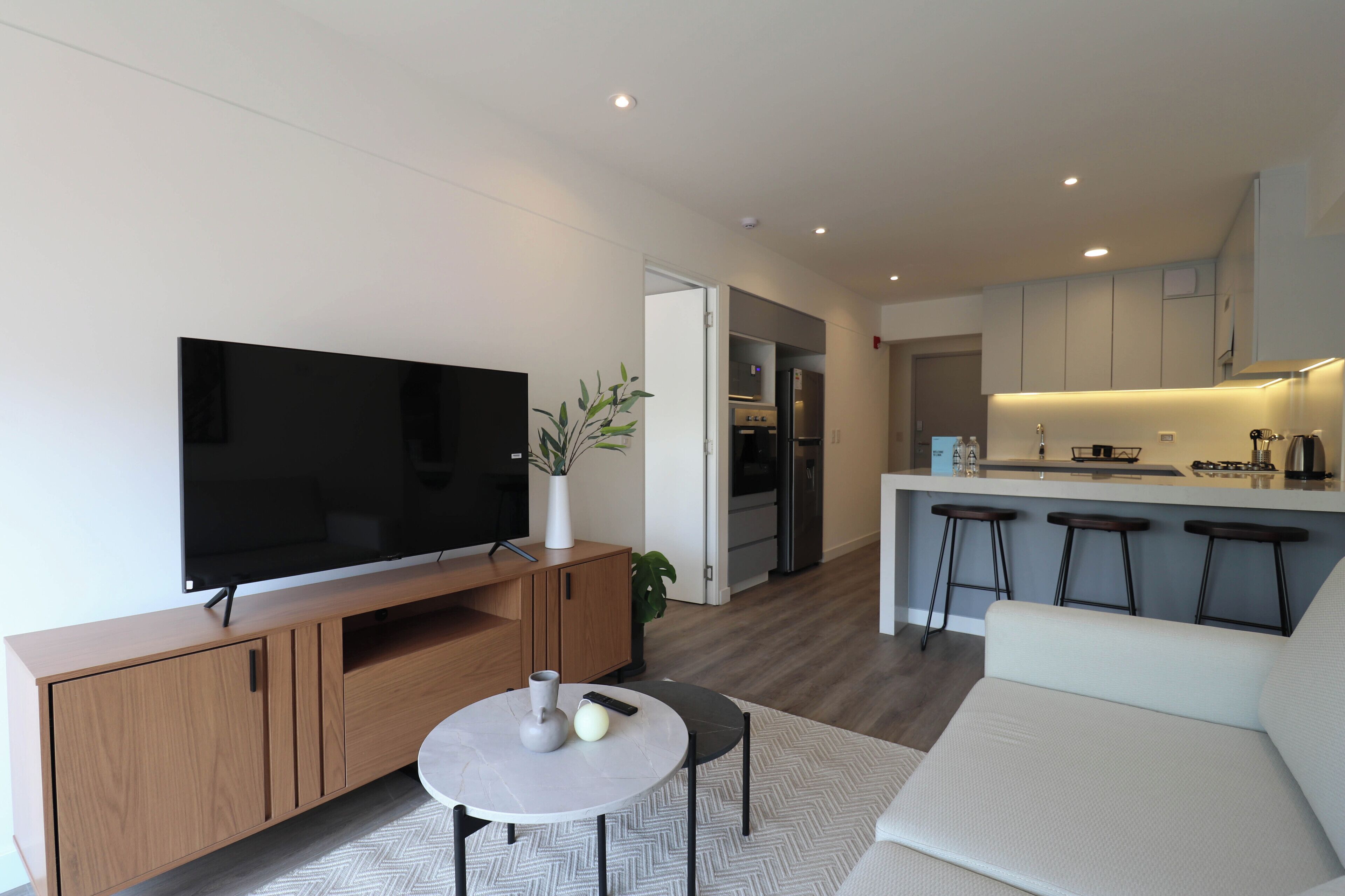 A modern living room with a flat-screen TV, a coffee table, and a sofa. There is a kitchen area with a bar and stools.
