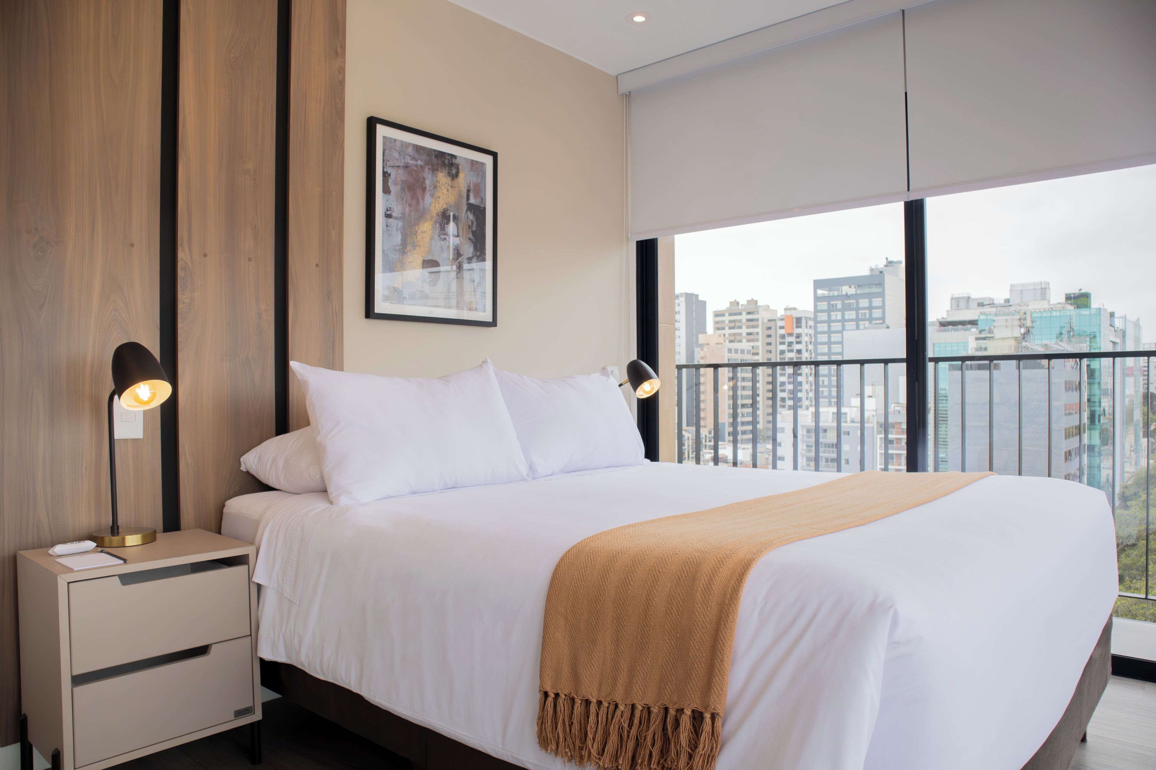 A modern hotel room with a large bed, a nightstand with a lamp, and a city view through large windows.