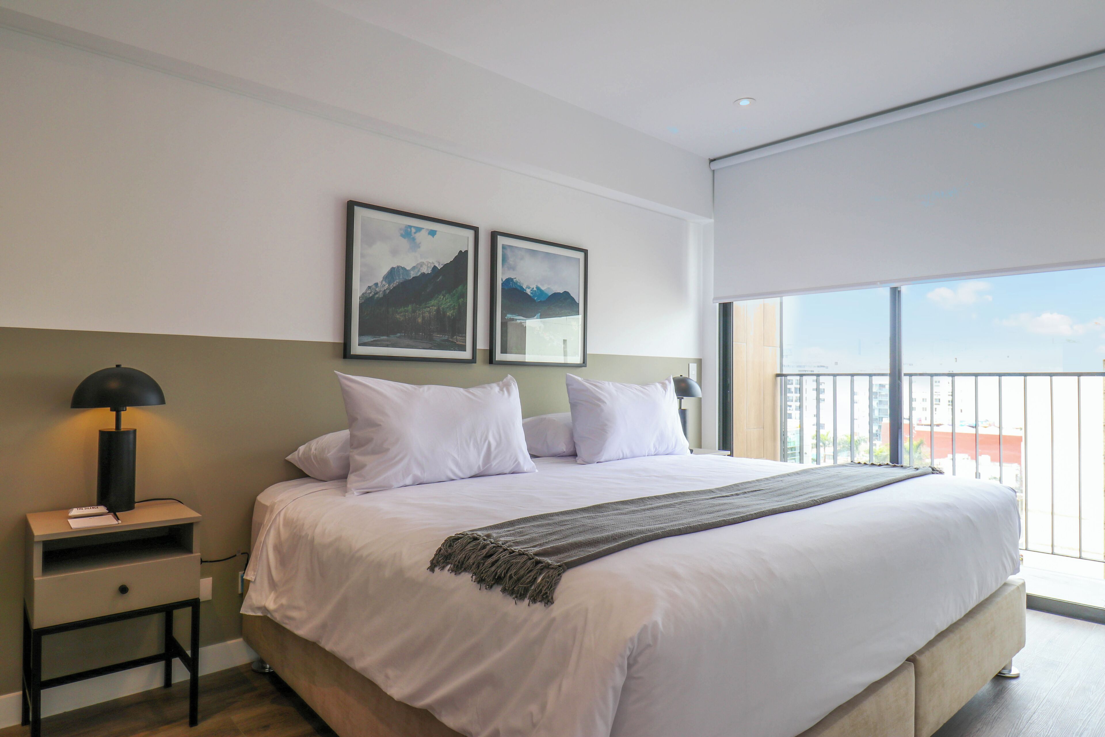 A bedroom with a large bed, a nightstand, two framed pictures on the wall, and a balcony with a view.