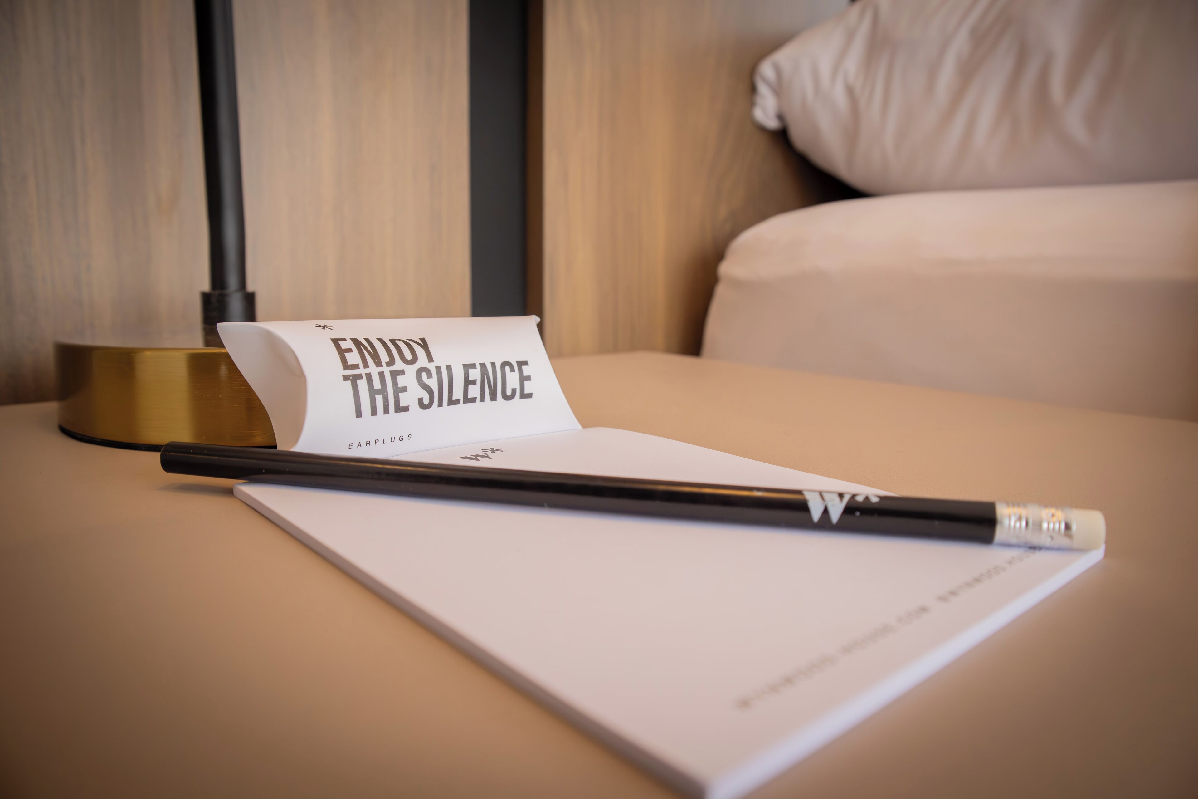 Un llit amb una almofada blanca i un llençol blanc, una ploma negra i un tros de paper amb la inscripció 'ENJOY THE SILENCE' sobre una taula de fusta.