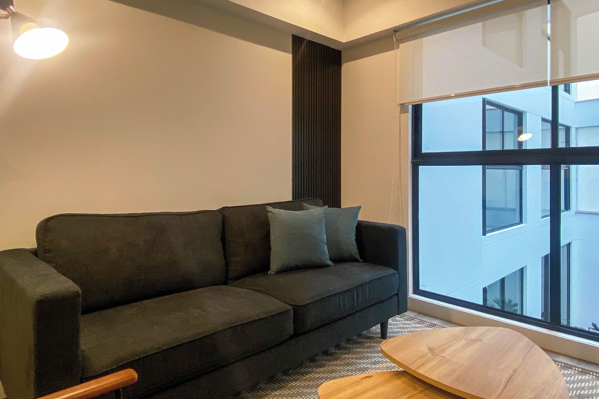 A modern living room with a dark grey sofa, a wooden coffee table, and a large window offering a view of a building.