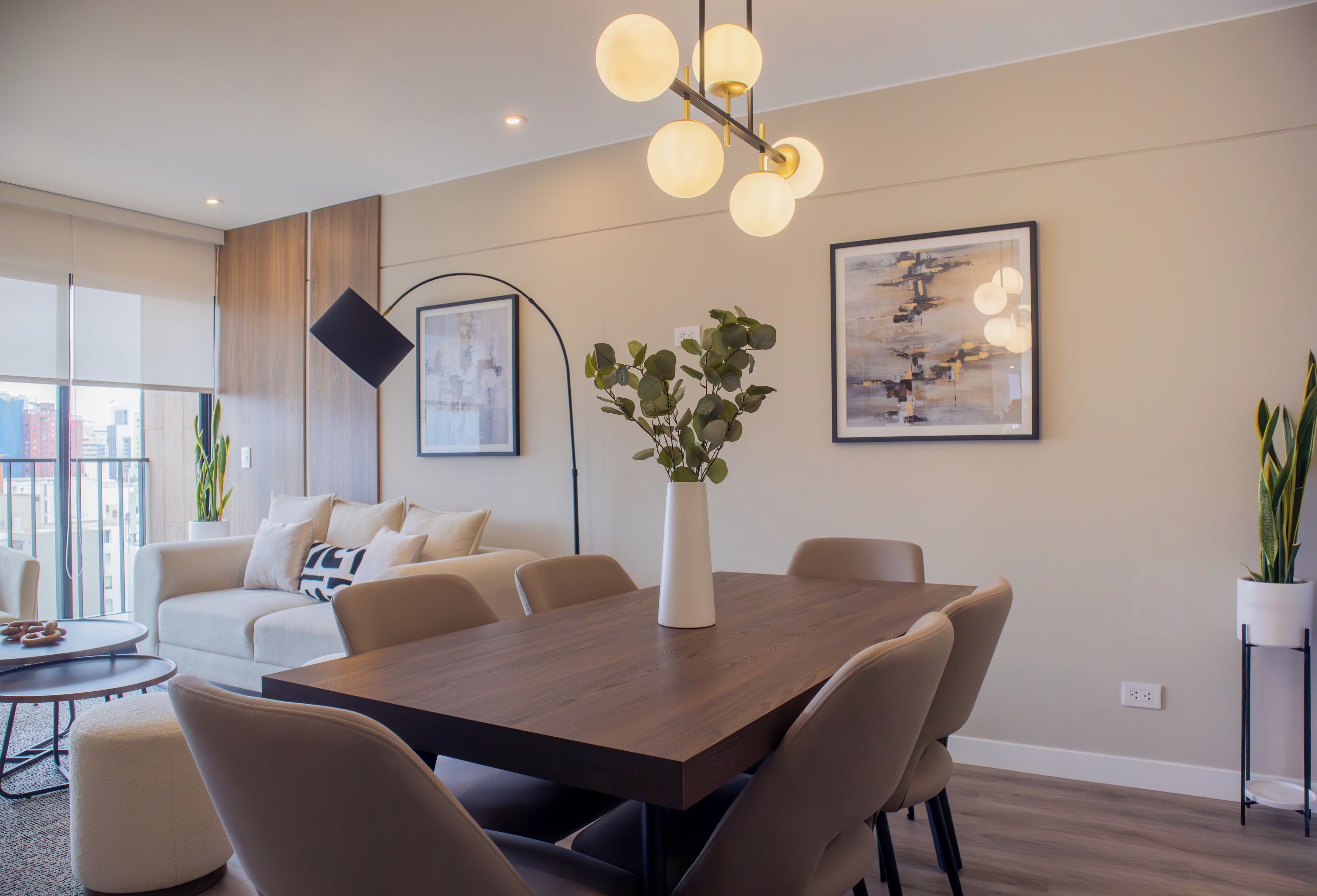 Una sala moderna con comedor, que incluye una mesa de madera, sofás color beige y un ventanal con vistas a la ciudad.